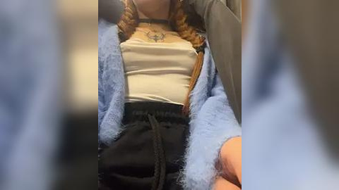 Media: Video of a person wearing a light blue fuzzy robe over a white tank top and black pants, with braided hair visible, seated on a car seat.