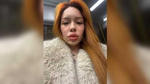 Media: Video of a young woman with long, vibrant orange hair, wearing a light fur coat, pouting, indoors with a blurred background.