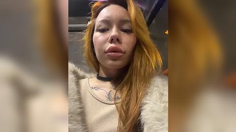 Media: Video of a young woman with long, straight, orange hair, wearing a beige sweater, a black choker, and a fur-lined jacket. She has a neutral expression and a slight pout.