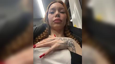 Media: Video of a Caucasian woman with light skin and long, curly auburn hair. She has a tattoo on her right hand and is wearing a black top. Background shows a blurred interior with a white curtain.