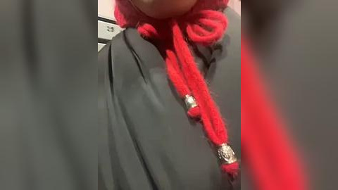 Media: Video of a person wearing a grey coat with a red, knitted scarf featuring a silver pendant, partially obscured by out-of-focus people in the background.