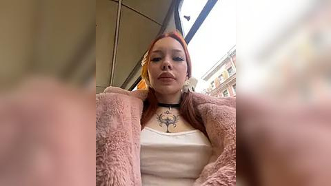 Media: Video of a young woman with red hair, wearing a white top and pink fuzzy coat, sitting in a bus. Her neck has a tattoo. Background shows urban buildings and a bus window.