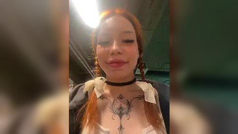 Media: Video of a young woman with long red hair, light skin, and tattoos on her chest, wearing a black choker, a white bra, and a dark jacket, indoors with a blurry background.