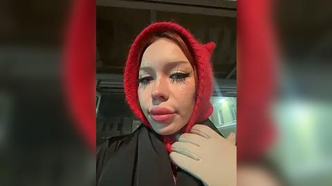 Media: Video of a fair-skinned woman with red hair, wearing a red devil hat, black top, and red lipstick, with a blurred background of a public transit bus.