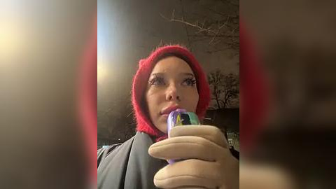 Media: Video of a woman with fair skin and a red beanie, wearing a gray coat, holding a colorful lollipop. Background shows blurry trees and a dimly lit scene.