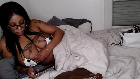 Media: A video of a dark-skinned woman with long black hair, glasses, and tattoos, breastfeeding while wearing a white bra, lying in a messy bed with white sheets and pillows.