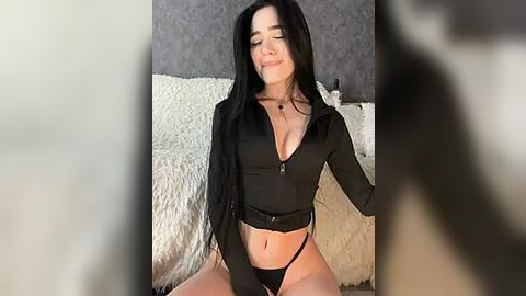 Media: A video of a young woman with long black hair, fair skin, and a slender physique, wearing a black, low-cut crop top and matching thong, sitting on a white fluffy rug in a dimly lit room.