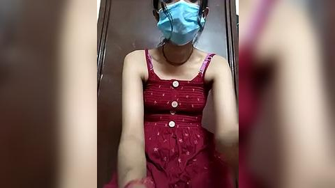 Media: A video of a woman in a red, patterned sundress with buttons, wearing a blue face mask, standing in a dimly lit elevator.