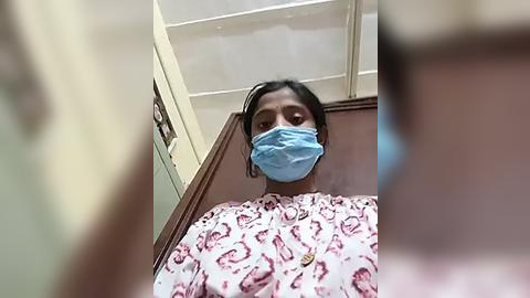 Media: Video of a person with a medium brown complexion, wearing a blue surgical mask, blue scrubs with a floral pattern, and a necklace, standing in a dimly lit room with white ceiling tiles.