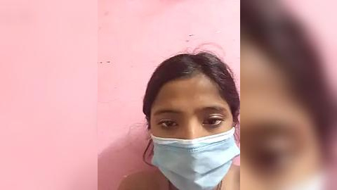 Media: Video of a young woman with dark skin and long black hair, wearing a blue surgical mask and a pink top, standing in front of a pink wall.