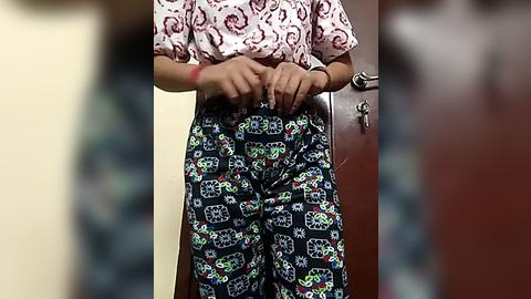 Media: Video of a person adjusting a colorful, patterned pair of pants with cartoon characters, wearing a white blouse with red swirls. Background shows a wooden door.