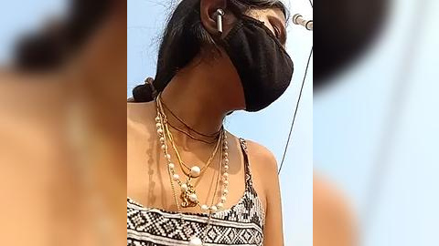 Media: Video of a woman with dark hair, wearing a black face mask, white and black patterned dress, and multiple gold necklaces, blurred background.