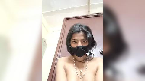 Media: A video of a young South Asian woman with shoulder-length black hair, wearing a black mask and a gold necklace, standing in front of a closed brown door in a dimly lit room.