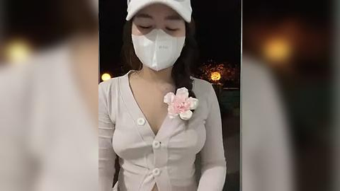 Media: Video of an Asian woman with long black hair, wearing a white surgical mask, a white cap, and a beige cardigan with a pink flower brooch, standing in a dimly lit outdoor setting at night.