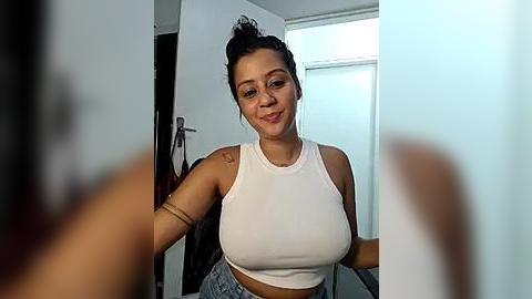 Media: Video of a Latina woman with medium skin tone and medium build, wearing a white tank top and high-waisted jeans, standing in a modern, minimalist room with white walls and a window.