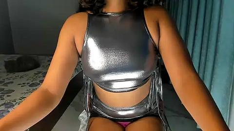 Media: Video of a woman with medium brown skin wearing a shiny, metallic silver crop top and high-waisted shorts, seated on a bed with a floral-patterned duvet.