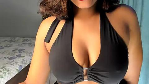 Media: Video of a woman with medium-dark skin, wearing a black halter top, revealing ample cleavage, sitting on a bed with floral-patterned sheets.