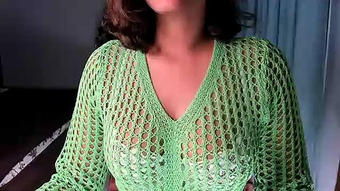 Media: Video of a woman with shoulder-length brown hair, wearing a see-through, green crochet top, revealing her breasts underneath, in a dimly lit room with a light blue curtain.