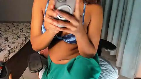 Media: Video of a woman in a blue bra, green skirt, holding a camera, sitting on a bed with floral sheets, in a dimly lit bedroom.