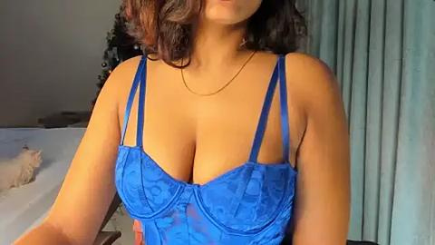 Media: Video of a woman with medium brown skin and curly brown hair, wearing a revealing, blue lace bra, indoors near a bed and teal curtains.