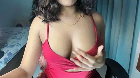 Media: A video shows a woman with medium brown skin, curly dark hair, wearing a low-cut red dress, revealing cleavage. She's indoors, near a bed with patterned bedding and light blue curtains.