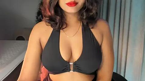 Media: Video of a woman with medium brown skin, wavy dark hair, and full red lips, wearing a black halter top with a zipper accentuating her cleavage, sitting on a bed in a dimly lit room with light blue curtains.