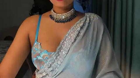 Media: A video of a woman with light brown skin, wearing a sheer blue saree with intricate embroidery, a matching turquoise bra, and a silver choker necklace.