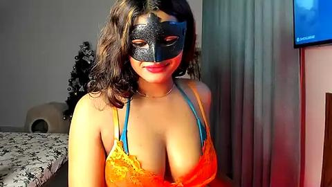 Media: Video of a woman with medium skin tone, wearing a black lace mask and orange lace bra, in a bedroom with a bed, Christmas tree, and TV.