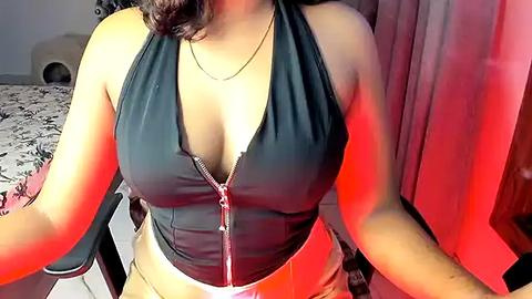 Media: Video of a woman with light brown skin, wearing a black halter top that accentuates her large breasts, sitting in a dimly lit room with a red glow.