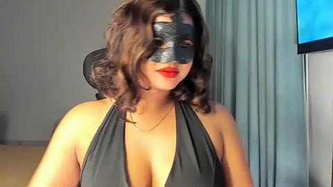 Media: Video of a woman with medium brown skin, wavy brown hair, and a black mask covering her eyes, wearing a black halter dress. Background features a TV screen and light grey curtains.