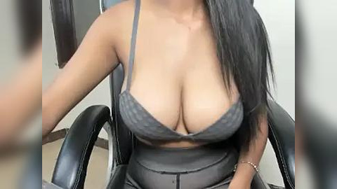 Media: Video of a light-skinned woman with long black hair, wearing a sheer black lace bra revealing ample cleavage, sitting in a black leather office chair, blurred background.