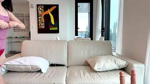 Media: Video of a modern, minimalist living room featuring a light beige sofa with two white pillows, a woman wearing a pink top, and a colorful abstract painting on the wall.