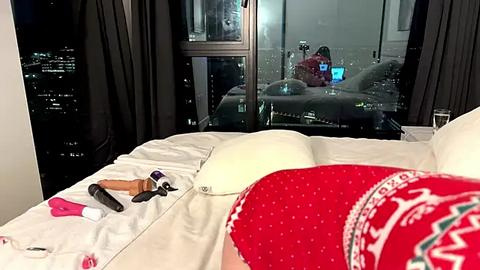 Media: Video of a cozy bedroom with a large window showing the nighttime cityscape. A red knitted hat and a blanket with a white pattern are visible. Makeup brushes and other personal items are scattered on the bed.