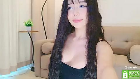 Media: Video of an East Asian woman with long, wavy black hair, light skin, and a slender physique, wearing a black strapless top. She stands in a modern living room with beige furniture and a green plant.