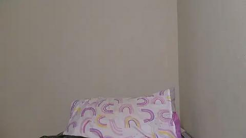 Media: Video of a minimalist bedroom corner with a white wall. A pillow with pastel rainbow stripes and a purple pillowcase lies on a bed, partially visible. The room is sparsely decorated, emphasizing simplicity and calmness.