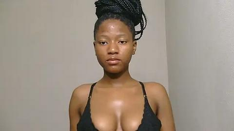 Media: Video of a dark-skinned woman with a high ponytail, wearing a black bra, against a plain beige wall. Her expression is neutral, and her breasts are prominently displayed.