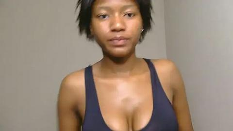 Media: Video of a young, African-American woman with a short, black hairstyle, wearing a low-cut, dark blue tank top, standing against a plain beige wall, giving a neutral expression.