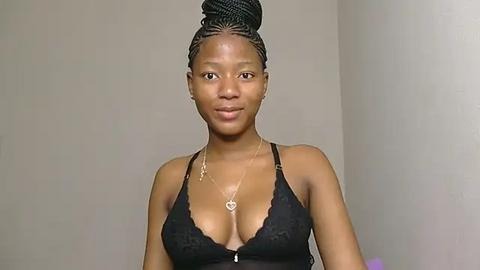 Media: Video of a slender, dark-skinned woman with a medium-sized bust wearing a black lace bralette. Her hair is styled in a high bun. She stands against a plain beige wall.