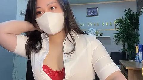 Media: Video of an East Asian woman with long black hair, wearing a white blouse, red lace bra, white mask, and black face shield, posing indoors with a desk and plants in the background.