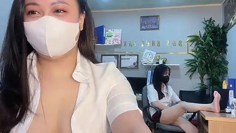 Media: Video of a woman in a white mask, white shirt, and black shorts, sitting at a desk in an office, with a green plant and certificates on the wall.