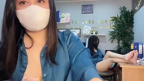 Media: Video of a smiling woman in a blue shirt and face mask, with another woman in a similar shirt lying on a desk in a brightly lit office.