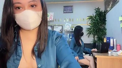 Media: Video of a woman with a mask, blue shirt, sitting in an office with plants, desks, and framed photos on the wall.