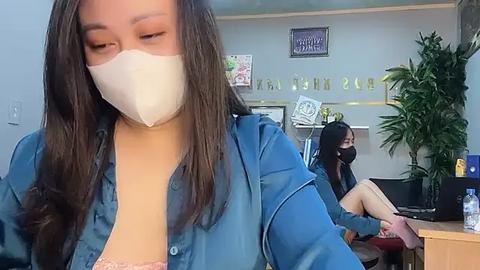 Media: Video of an Asian woman with long dark hair wearing a white mask, blue shirt, and pink bra. Background shows another Asian woman with long dark hair, also masked, seated at a desk.