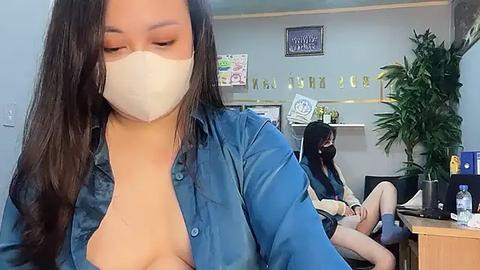 Media: Video of an Asian woman in a blue shirt, wearing a face mask, exposing her bare chest. Another Asian woman, also masked, sits in the background.