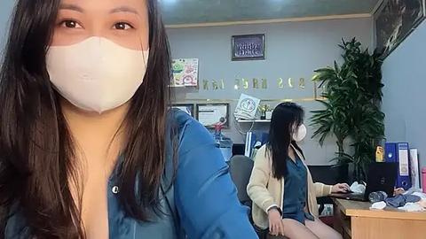 Media: Video of an office scene with a woman wearing a face mask, blue shirt, and long brown hair, sitting next to a woman in a yellow jacket and long black hair, typing on a computer.