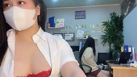 Media: Video of a woman in a white shirt with a red lace bra, white mask, and long dark hair, in an office with green plants, certificates, and a desk.