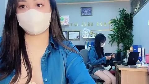 Media: Video of two East Asian women wearing blue scrubs and masks, seated at desks in a medical office; one woman is typing, the other looks at a computer.