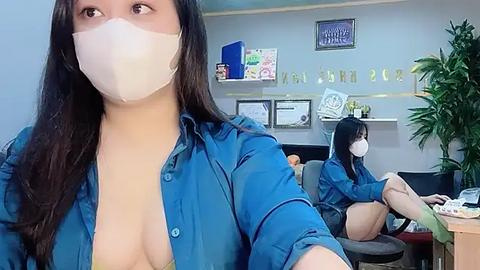 Media: Video of two Asian women wearing blue shirts and face masks, sitting in an office with plants and framed certificates on the wall.