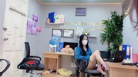 Media: Video of an office room with a woman wearing a face mask, blue sweater, and pink shorts sitting on a chair, surrounded by office supplies and plants.