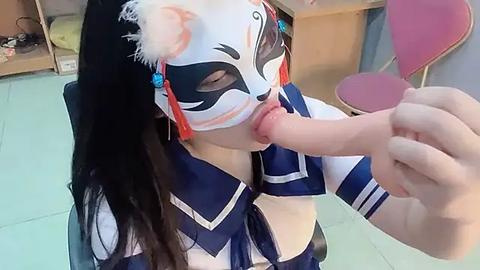 Media: Video of an Asian woman in a white fox mask, blue and white sailor outfit, performing oral sex on a circumcised penis in a modern room with wooden furniture and pink chairs.